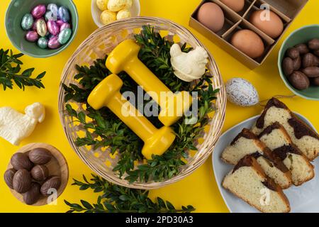 Gym dumbbells in traditional Easter Paschal wicker basket. Healthy diet choice concept. Workout flat lay with chocolate candies, cake, eggs, boxwood. Stock Photo