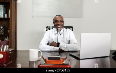 A mature African man sits at a table indoors, confidently crossing his arms as he uses his laptop for corporate business. Stock Photo