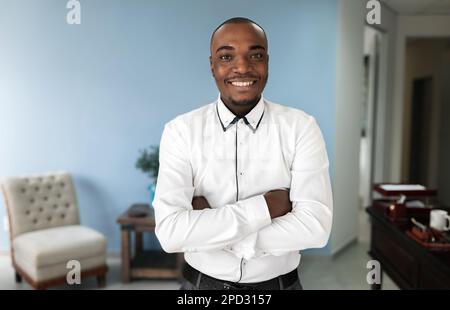 A cheerful, African man stands in an indoor office setting with arms crossed and a smiling face as he looks directly at the camera. Stock Photo