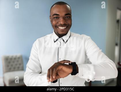 A cheerful adult African business man stands wearing a smart watch, looking at the camera with happiness. He is dressed for his occupation and enjoyin Stock Photo