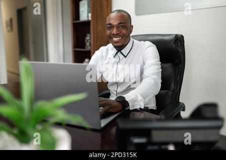 A smiling African businessman sits in his office, looking at the camera while working on a laptop connected to wireless technology. Stock Photo