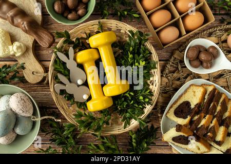 Gym dumbbells in traditional Easter wicker basket. Healthy diet choice concept. Fitness workout flat lay with chocolate candies, cake, eggs, boxwood. Stock Photo