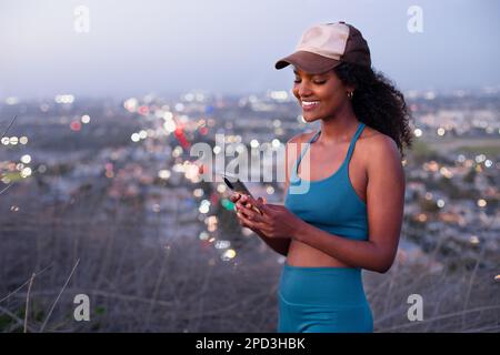 Young woman in blue sports clothing & baseball cap using smartphone in park Stock Photo