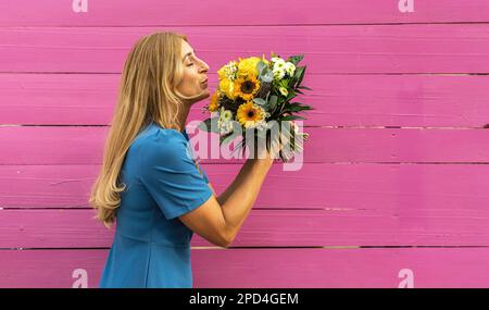 woman holding and smells on a bunch of spring flowers, dressed in blue clothes in front of wooden pink background Stock Photo