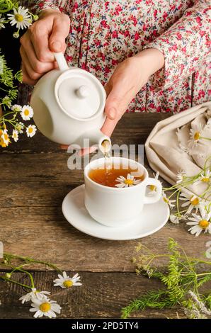 Woman pouring chamomile herbal tea on dark wooden background. Stock Photo