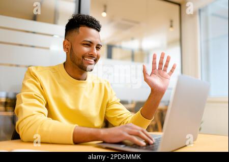 Male indian student or freelancer man using laptop for virtuel connection, smiling guy involved virtual meeting sitting at the office desk, cheerful man waving, making video call, taking online course Stock Photo