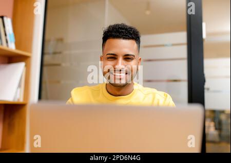 Portrait of young indian man freelancer or student in casual yellow pullover using laptop sitting in modern office or library, male office employee typing messages, web surfing, front view Stock Photo