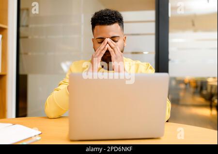 Fatigued Indian man using laptop for working. Male employee in casual wear touching and massaging the bridge of the nose, feeling tired and burnout, while sitting at desk in office with eyes closed Stock Photo