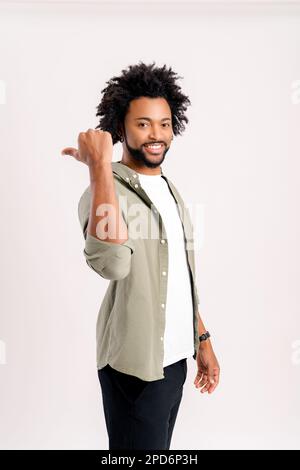 Vertical view of the joyful curly man presenting advertising area with big finger, holding empty place for commercial. Indoor studio shot isolated on white background Stock Photo