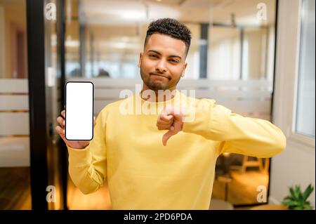 Portrait of young indian man showing smartphone with empty screen and thumbs down, expressing disapproval, criticizing and disagreeing with suggestion, standing in the modern office Stock Photo