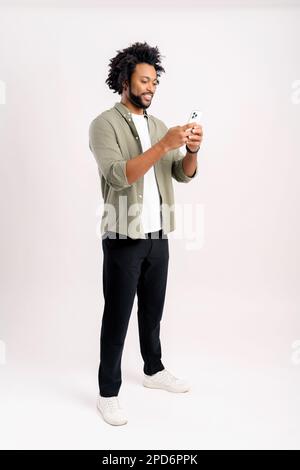Portrait of cheerful young black guy sending message on mobile phone against white background. Happy African-American man using smartphone, enjoying chatting online, messaging in social networks Stock Photo