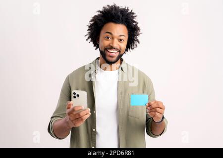 Cheerful middle eastern guy is using smartphone and credit card for shopping online. Happy man is ordering food online, male paying for long-awaited purchase on the mobile phone, isolated on white Stock Photo