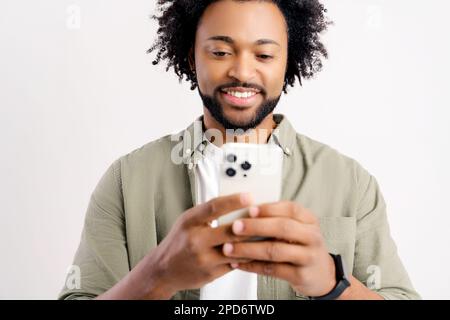 Portrait of cheerful young black guy sending message on mobile phone over white background. Happy African-American man using smartphone, enjoying chatting online, messaging in social networks Stock Photo