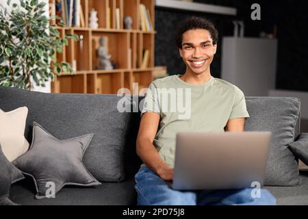 Handsome young multiracial man with beard sitting on the sofa with laptop at home, searching in the internet, watching film or serial, remote meeting, on the weekend, relaxed, listening performance Stock Photo