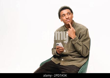 Thoughtful young african-american man sitting on armchair with smartphone and touching chin, looking for new solution of tasks, choosing, isolated over white background Stock Photo