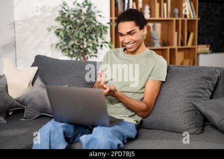 Young good-looking man is using a laptop for video connection, making real call, long-waited meeting, gesturing, looking at the computer screen, sitting on the sofa and holding stylish laptop on laps Stock Photo