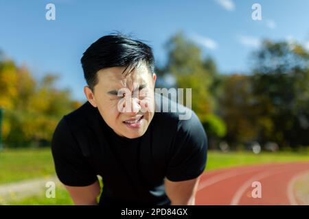 Close-up of sportsman having severe headache and dizziness, asian man in stadium doing fitness and active exercise, male runner in sportswear overworked by training on sunny day. Stock Photo