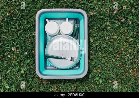 Washing up bowl filled with the washed outdoor dishes, plates, cups and cutlery put on grass on camping Stock Photo