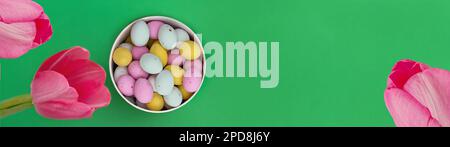 Banner made from Multi Colored chocolate eggs in box and pink tulips on green background. Egg colors: pink, yellow and white. Copy space. Ideal for spring themes, use in marketing materials, packaging Stock Photo