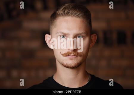 Fashion young man model with stylish hairdo, beard and 'handlebar' moustache looks confidently at camera on brick wall background. Barber fashion and beauty. Beauty hair salon, barbershop. Stock Photo