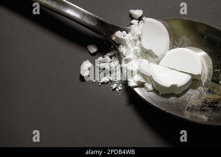 Pharmaceutical medicine drugs powder on a silver metal spoon. Smashed remedy, tablets, on top of a grey table in the background. Stock Photo