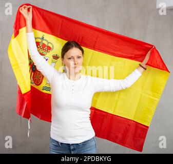 Unhappy sad middle-aged woman waving national flag of Spain while looking at camera at gray wall background Stock Photo