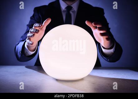 Businessman using glowing crystal ball to predict future at table, closeup. Fortune telling Stock Photo