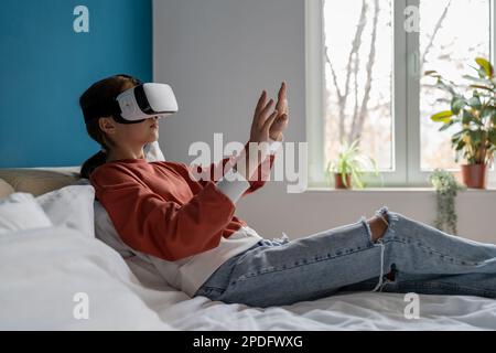 Teen girl using modern VR glasses while resting at home, touching things in metaverse Stock Photo