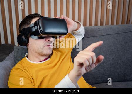 Smiling mature Adult male wearing VR glasses presses by his finger on virtual reality interface at home, game, technology, metaverse, viewing 360 vide Stock Photo
