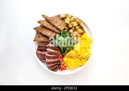 Tenderloin beef steak with scrambled eggs, rocket leaves and potatoes with a side of rye bread. Stock Photo