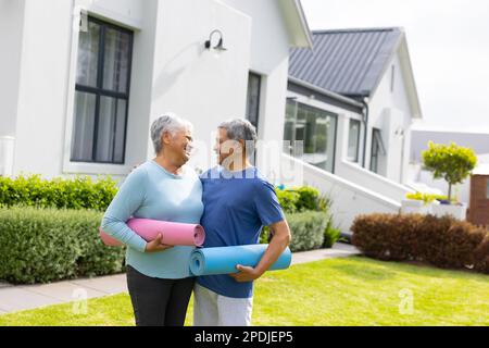 Romantic biracial senior couple with exercise mats looking at each other while standing over house Stock Photo