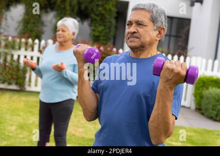 Biracial senior couple lifting dumbbells while standing and exercising in yard Stock Photo