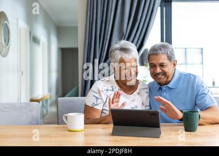 Smiling biracial senior couple waving hands while talking over video call on digital tablet at home Stock Photo