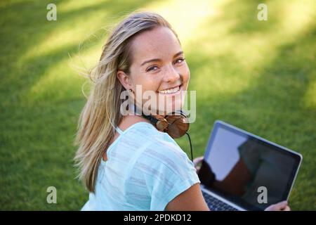 I have all the research I need at my fingertips. Portrait of a smiling young woman sitting on the grass in a park and using her lap top. Stock Photo
