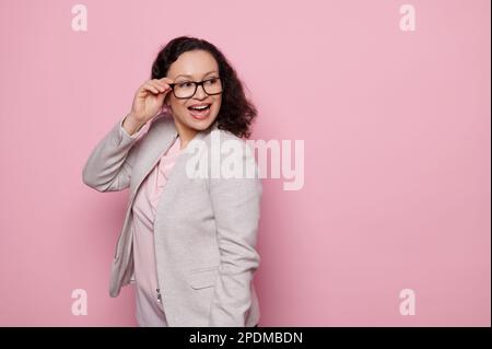 Charming dark-haired woman smiling, looking aside, wearing trendy glasses and elegant light gray suit, pink background Stock Photo