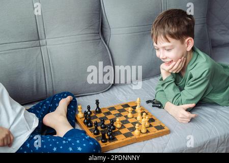 Boys play wooden chess lying on a gray sofa. Stock Photo