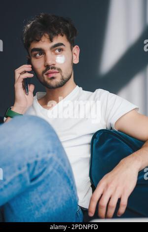 Young caucasian man sitting on sofa at home talking on smart phone in living room. Having conversation. Looking away. Home comfort. Satisfied person Stock Photo