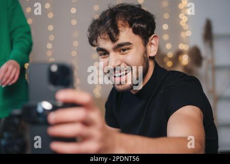 Portrait of positive young man filming tutorial video trend for social network, using cellphone on tripod in living room. Internet technology, digital Stock Photo