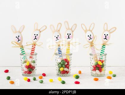 Handmade cute colorful bunnies made from wooden spoons in glasses with sweet candy eggs. Small gift or decor for Easter. Easy fun kids crafts concept. Stock Photo