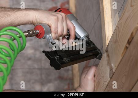 Unrecognizable cropped male hands using pneumatic nailer, nail gun with spiral cable for wooden timber board. Make layout on wooden plank. Clipping Stock Photo