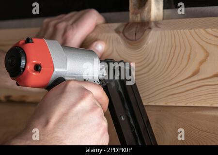 Close up cropped carpenter male hands using pneumatic nailer, stapler gun for wooden timber plank. Making layout on wooden plank. Builder handwork and Stock Photo
