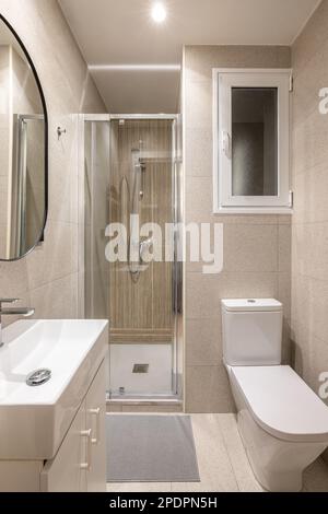 Modern compact beige tiled bathroom with glass walk-in shower, sink and mirror and white toilet with window. Concept of a tiny bathroom in a hotel or Stock Photo