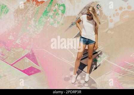 oung attractive and beautiful girl is standing on a graffiti wall background. Woman is wearing white, empty basic t-shirt without logo. Vertical mock- Stock Photo