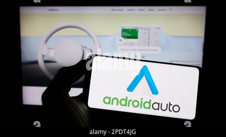 Person holding cellphone with logo of mobile app Android Auto (Google) on screen in front of webpage. Focus on phone display. Stock Photo
