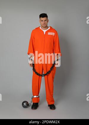 Prisoner in jumpsuit with chained hands and metal ball on grey background Stock Photo