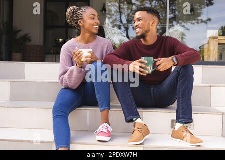 Happy african american couple sitting on stairs in garden, drinking coffee Stock Photo