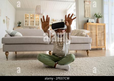 Happy African American kid boy in virtual reality glasses exploring metaverse at home Stock Photo