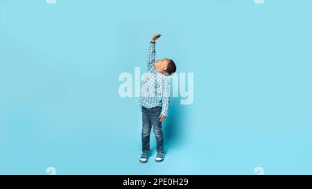 Little african american boy wishes he were taller, raising his hand to show how tall he wants to grow, blue background Stock Photo