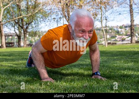 Portrait of a senior man exercising, doing push ups in the park. An elderly man in good shape and health with strong arms. Stock Photo