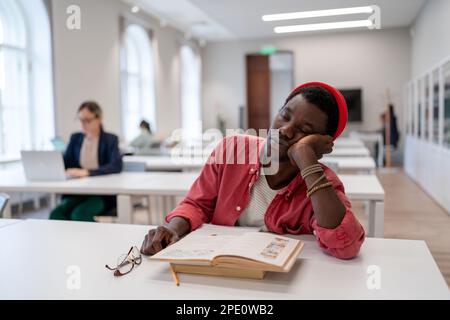 Sleepy lazy African American man falls asleep sits in library with head resting on desk with books Stock Photo